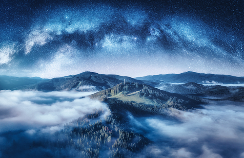 Milky Way arch and mountains in low clouds at starry night in summer. Landscape with sky with stars, arched Milky Way, trees on the hill in fog, mountain peaks. Space and galaxy. Aerial view. Nature