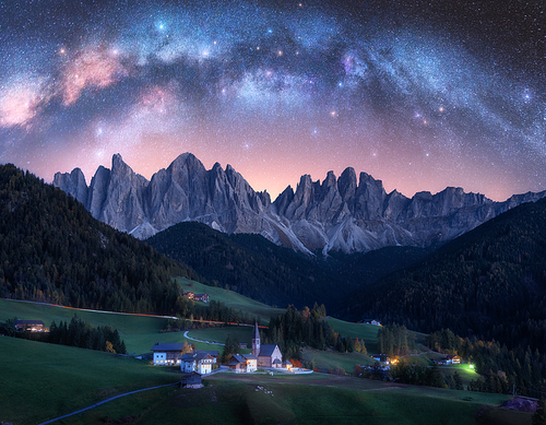Santa Maddalena and acrhed Milky Way at night in summer in Italy. Starry sky with milky way arch over St. Magdalena and mountains. Village with houses, church, green meadows, trees, rocks. Space