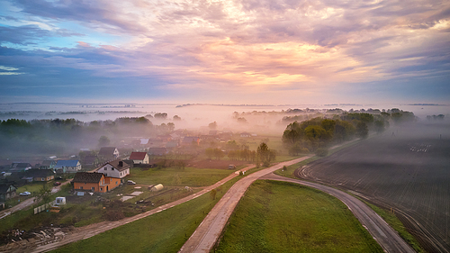 Aerial view of village, rural dirt road and trees covered by fog. Early misty morning sunrise panorama. Spring summer fields. Rainy overcast moody weather. Belarus, Minsk region