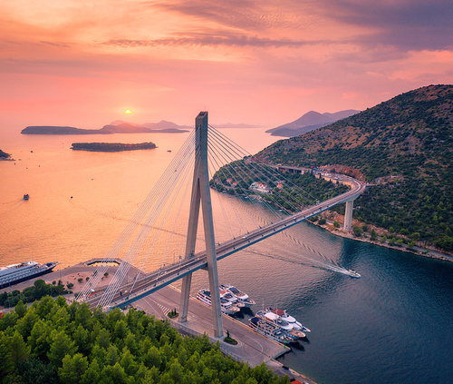 Aerial view of beautiful modern bridge at sunset in summer. Dubrovnik, Croatia. Top view of road, boats, yachts, trees. Summer landscape with harbor, mountain, islands, highway, blue sea and red sky