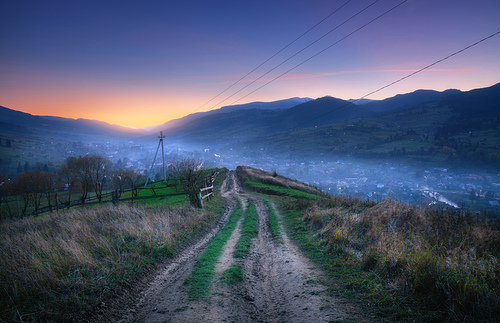 Mountain dirt road at beautiful sunset in summer. Colorful landscape with road, village in fog, blue sky with sunlight, mountains, trees at night. Trail on the hill. Travel and nature. Scenery