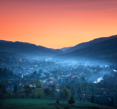 Mountain village in fog at beautiful sunset in spring. Colorful rural landscape with buildings in low clouds, mountains, trees and orange sky at night in Ukraine. Foggy town at twilight. Travel