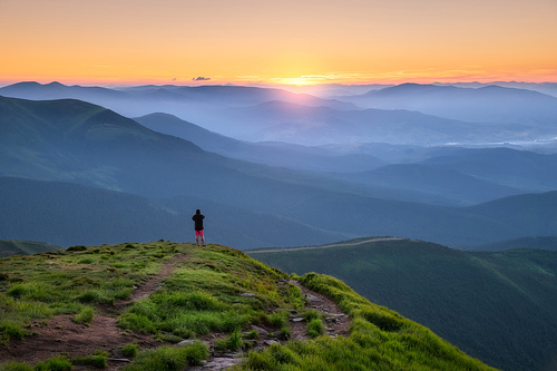 Sporty man on the mountain peak looking on mountain valley with sunbeams at colorful sunset in summer in Europe. Landscape with traveler, foggy hills, forest in fall, amazing sky and sunlight