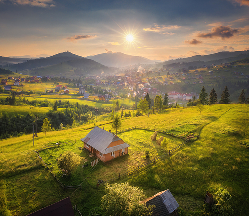 Aerial view of beautiful village in Carpathian mountains at sunset in summer. Colorful landscape with green meadows, houses with gardens, trees, sky with clouds. Top view of mountain countryside