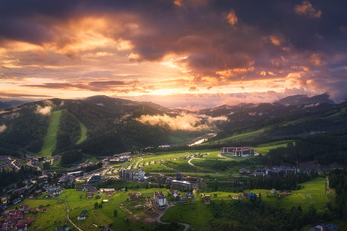 Aerial view of beautiful village in Carpathian mountains at sunset in summer. Colorful landscape with green meadows, houses, trees, dramatic sky. Top view of mountain countryside. Bukovel, Ukraine