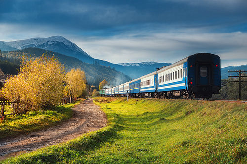 Moving train in mountains at sunset in autumn. Industrial landscape with passenger speed train on railroad, dirt road, snowy rocks, orange trees, green grass, purple sky in fall. Rural railway station