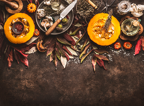 Autumn seasonal food background with halved pumpkin, knife , herbs and spices on dark rustic background with fall leaves. Top view. Border