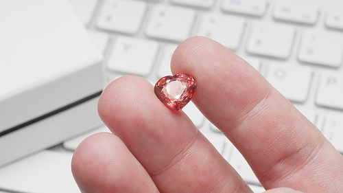 Heart shaped tourmaline gem in hand of gemologist or jeweller. Online jewelry shopping for birthday, Valentine Day or other holiday. Male hand holding red tourmaline, macro shot with box and keyboard.