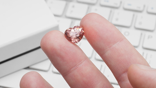 Heart shaped tourmaline gem in hand of gemologist or jeweller. Online jewelry shopping for birthday, Valentine Day, other holiday. Male hand holding peach tourmaline, macro shot with box and keyboard
