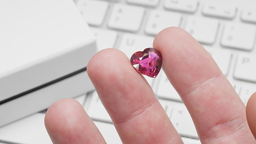 Heart shaped tourmaline gem in hand of gemologist or jeweller. Online jewelry shopping for birthday, Valentine Day or other holiday. Male hand holding red tourmaline, macro shot with box and keyboard.