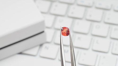 Orange spinel gem in tweezers. Online jewelry shopping for birthday, Valentine Day, other holiday. Gemologist or jeweller holding red spinel, macro shot with box and keyboard