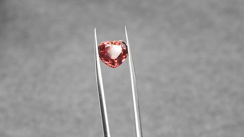 Red heart shaped tourmaline gem in tweezers. Jewelry shopping for birthday, Valentine Day, other holiday. Gemologist or jeweller holding red tourmaline, macro shot on gray background