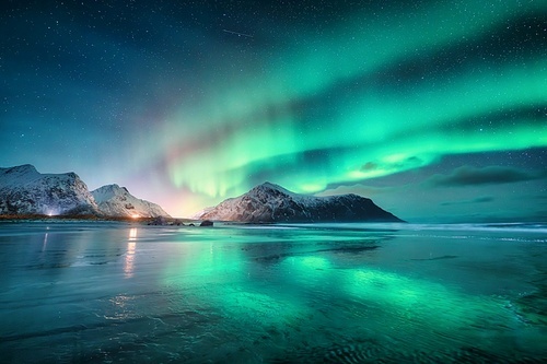 Northern Lights and sandy beach at starry winter night. Lofoten islands, Norway. Beautiful Aurora borealis. Sky with polar lights. Landscape with aurora, sea, sky reflection in water, snowy mountains