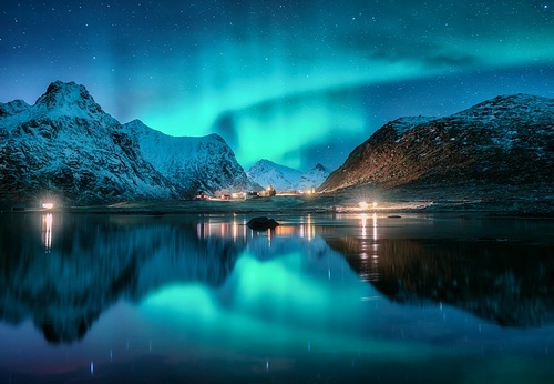 Aurora borealis, snowy mountains, sea, fjord, reflection in water, street lights at starry winter night. Lofoten, Norway. Northern lights. Landscape with polar lights, snowy rocks, sky with stars