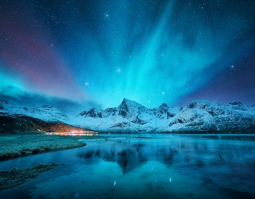 Northern lights over the snowy mountains, frozen sea, reflection in water at winter night in Lofoten, Norway. Aurora borealis and snowy rocks. Landscape with polar lights, starry sky and fjord. Nature