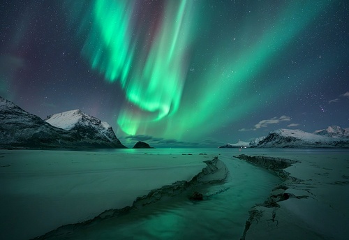 Northern Lights, snowy mountains, sandy beach, creek at starry winter night. Lofoten islands, Norway. Amazing Aurora borealis. Sky with polar lights. Landscape with aurora, sea, stream, rocks in snow