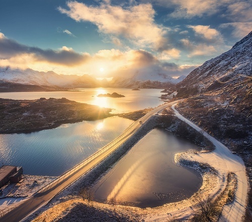 Aerial view of bridge, sea and snowy mountains at sunrise in Lofoten Islands, Norway. Landscape with beautiful road, water, rocks, blue sky with clouds and golden sunlight. Top drone view. Nature