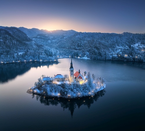 Aerial view of church on snowy island on the Bled Lake, Slovenia at winter night. Top drone view of chapel, alpine mountains, illumination, trees in snow, lights, reflection in water at sunset. Dusk