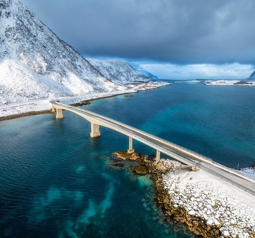 Aerial view of bridge over the sea and snowy mountains in Lofoten Islands, Norway. Top drone view of bridge in cold winter day. Wintry landscape with blue water, rocks in snow, road, cloudy sky.