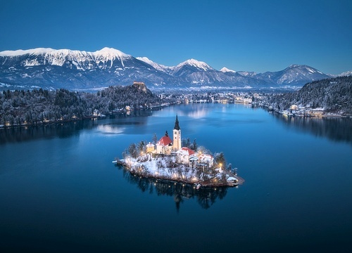 Aerial view of church on snowy island on the Bled Lake, Slovenia at winter night. Top drone view of chapel, alpine mountains, illumination, trees in snow, lights, reflection in water at sunset. Dusk