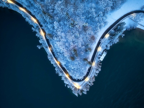 Aerial view of road, snowy forest, illumination, lake, street lights at winter night. Top drone view of alpine countryside, road through the snowy pine trees at dusk. Bled lake, Slovenia. Travel