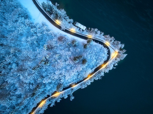 Aerial view of road, snowy forest, illumination, lake, street lights at winter night. Top drone view of alpine countryside, road through the snowy pine trees at dusk. Bled lake, Slovenia. Travel