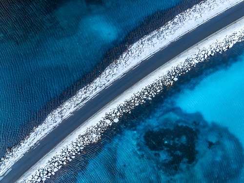 Aerial view of snowy road to the island and blue sea on both side. Bridge on snow and azure transparent water in winter. Landscape. Top drone view of road to the Henningsvaer, Lofoten islands, Norway