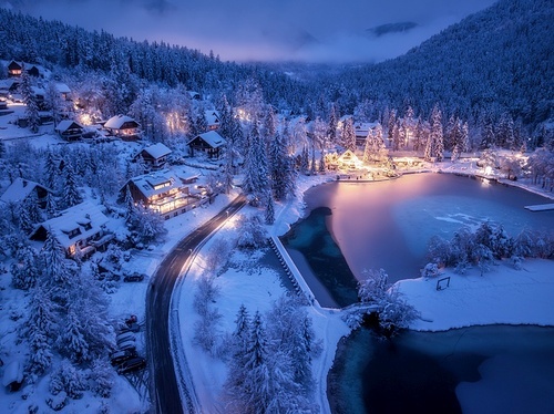 Aerial view of fairy alpine village in snow, road, forest, Jasna lake, houses, street lights at winter night. Top view of mountains, illumination, snowy pine trees at twilight. Kranjska Gora, Slovenia