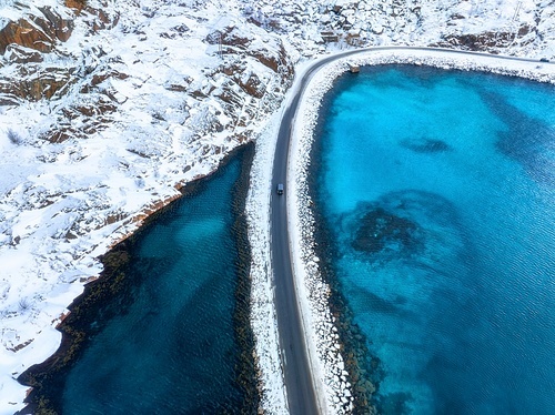 Aerial view of snowy road to the island and blue sea on both side. Bridge on snow and azure transparent water in winter. Landscape. Top drone view of road to the Henningsvaer, Lofoten islands, Norway