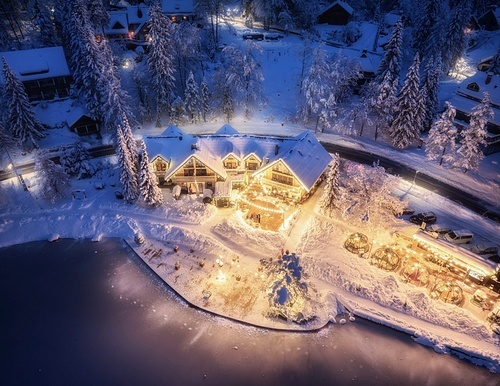 Aerial view of illuminated houses in fairy village in snow, forest, Jasna lake, street lights at winter night. Top view of alpine countryside, snowy pine trees at dusk. Kranjska Gora, Slovenia