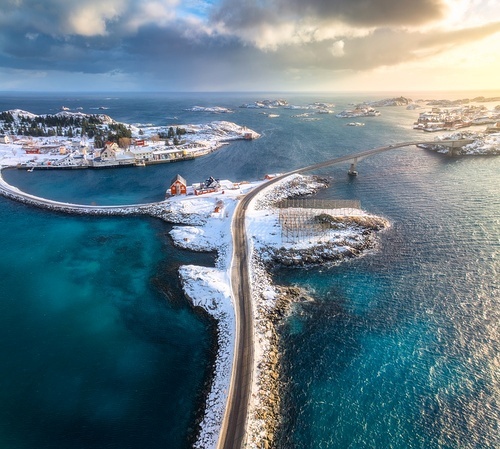 Aerial view of road and bridges, snowy rocks, islands with rorbu, sea, water, mountains, cloudy sky at sunset in winter. Beautiful north landscape. Top view of Henningsvaer, Lofoten islands, Norway