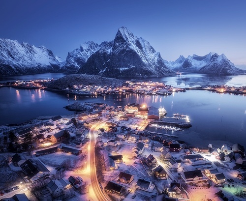 Aerial view of snowy village, islands, rorbu, city lights, blue sea, rocks and mountain at night in winter. Beautiful landscape with town, street illumination. Top view. Reine, Lofoten islands, Norway