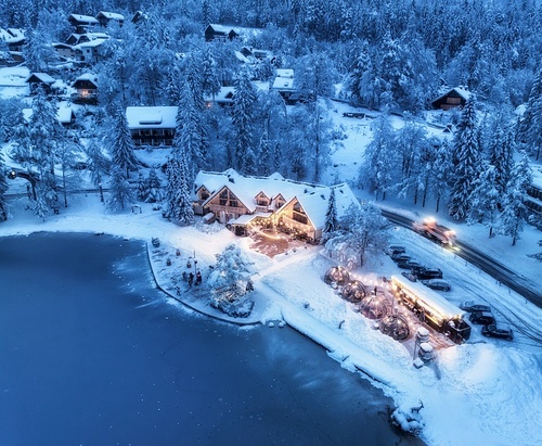 Aerial view of illuminated houses in fairy village in snow, forest, Jasna lake, street lights at winter night. Top view of alpine countryside, snowy pine trees, road at dusk. Kranjska Gora, Slovenia