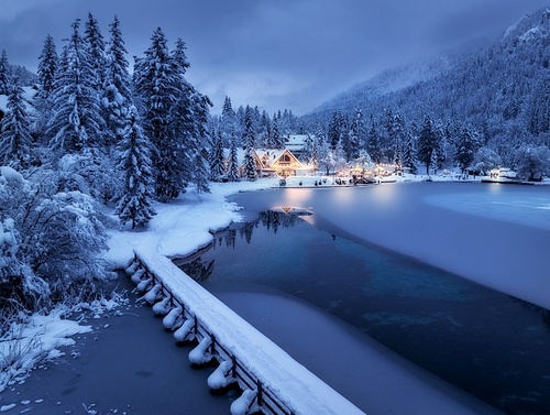 Aerial view of fairy alpine village in snow, bridge, forest, Jasna lake, houses, street lights at winter night. Top view of mountains, illumination, snowy pine trees at dusk. Kranjska Gora, Slovenia