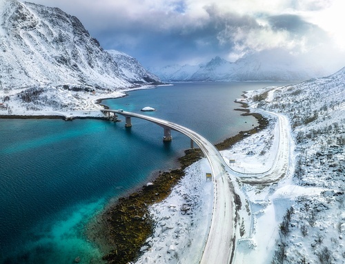 Aerial view of bridge over the sea and snowy mountains in Lofoten Islands, Norway. Top drone view of bridge in cold winter day. Wintry landscape with blue water, rocks in snow, road, cloudy sky.