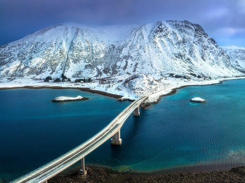 Aerial view of bridge over the sea and snowy mountains in Lofoten Islands, Norway. Top drone view of bridge in cold winter day. Wintry landscape with blue water, rocks in snow, road, cloudy sky.