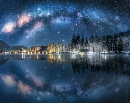 Milky Way arch reflected in water at winter starry night. Landscape with acrhed Milky Way reflected in Bled Lake, Slovenia. Sky with bright stars, snowy pine trees, street lights, illumination. Space