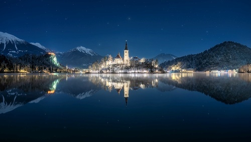 Beautiful church on the snowy island on the Bled Lake, Slovenia at winter starry night. Landscape with chapel, illumination, sky with stars, alpine mountains, trees in snow, reflection in water