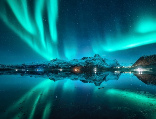 Northern lights, snowy mountains, sea, fjord, reflection in water, street lights at starry winter night. Lofoten, Norway. Aurora borealis. Scenery with bright polar lights, snowy rocks, sky with stars