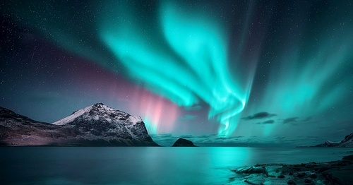 Northern Lights over the sea, snowy mountains at starry winter night. Aurora borealis in Lofoten islands, Norway. Sky with polar lights. Landscape with aurora, beach, sky, reflection in water. Space