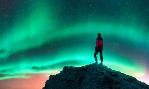 Northern lights and sporty woman on mountain peak at night. Aurora borealis and silhouette of a girl on top of rock. Landscape with polar lights. Starry sky with bright aurora. Travel background