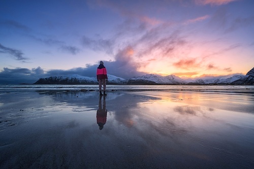 Woman on the sandy beach, snowy mountains and colorful sky at sunset in winter. Lofoten islands, Norway. Landscape with silhouette of a girl, sea, reflection in water, purple sky with pink clouds