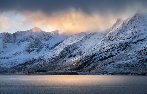 Snowy mountains in low clouds, sea bay, reflection in water at sunset in winter. Lofoten islands, Norway. Colorful landscape with rocks in snow, orange sky. Seashore. Nature background. Wintry scenery