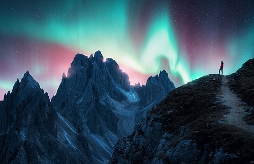 Northern lights and woman on mountain peak at night. Colorful aurora borealis and silhouette of a girl on mountain trail. Landscape with polar lights. Starry sky with bright aurora. Travel background