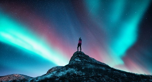 Northern lights and sporty woman on mountain peak at night. Aurora borealis and silhouette of a girl on top of rock. Landscape with polar lights. Starry sky with bright aurora. Travel background