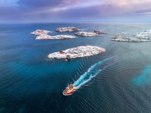 Aerial view of floating fishing boat in blue sea in snowy Henningsvaer village, Lofoten islands, Norway in winter. Top drone view of boat, mountains in snow, sea, cloudy sky, town, rorbuer and houses