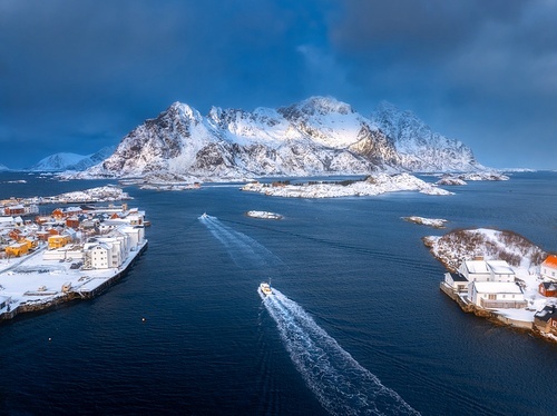 Aerial view of floating fishing boat in blue sea in snowy Henningsvaer village, Lofoten islands, Norway in winter. Top drone view of boat, mountains in snow, sea, cloudy sky, town, rorbuer and houses