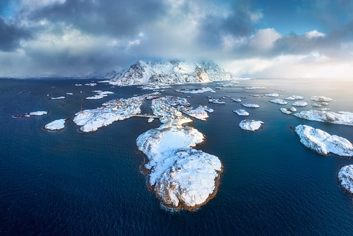 Aerial view of snowy Henningsvaer fishing village, Lofoten islands, Norway in winter at sunset. Top drone view of mountains in snow, sea, boats, cloudy sky, town, rorbuer and houses. Panorama. Scenery