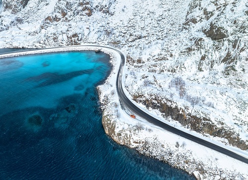 Aerial view of curved snowy road to the island and blue sea. Highway in snow and azure transparent water. Winter landscape. Top drone view of road in Lofoten islands, Norway. Transportation. Travel