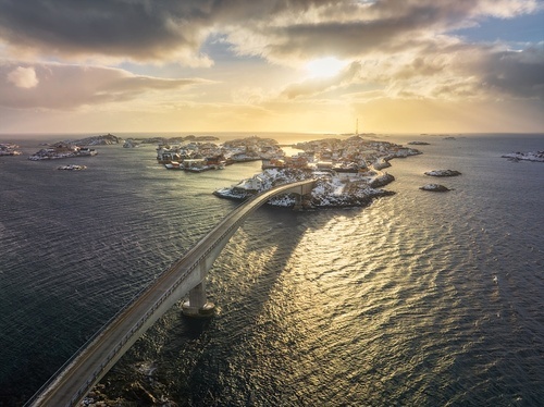 Aerial view of bridge, snowy rocks, islands with rorbu, sea, water, mountains, orange sky with clouds at sunset in winter. Beautiful landscape. Top view of road. Henningsvaer, Lofoten islands, Norway